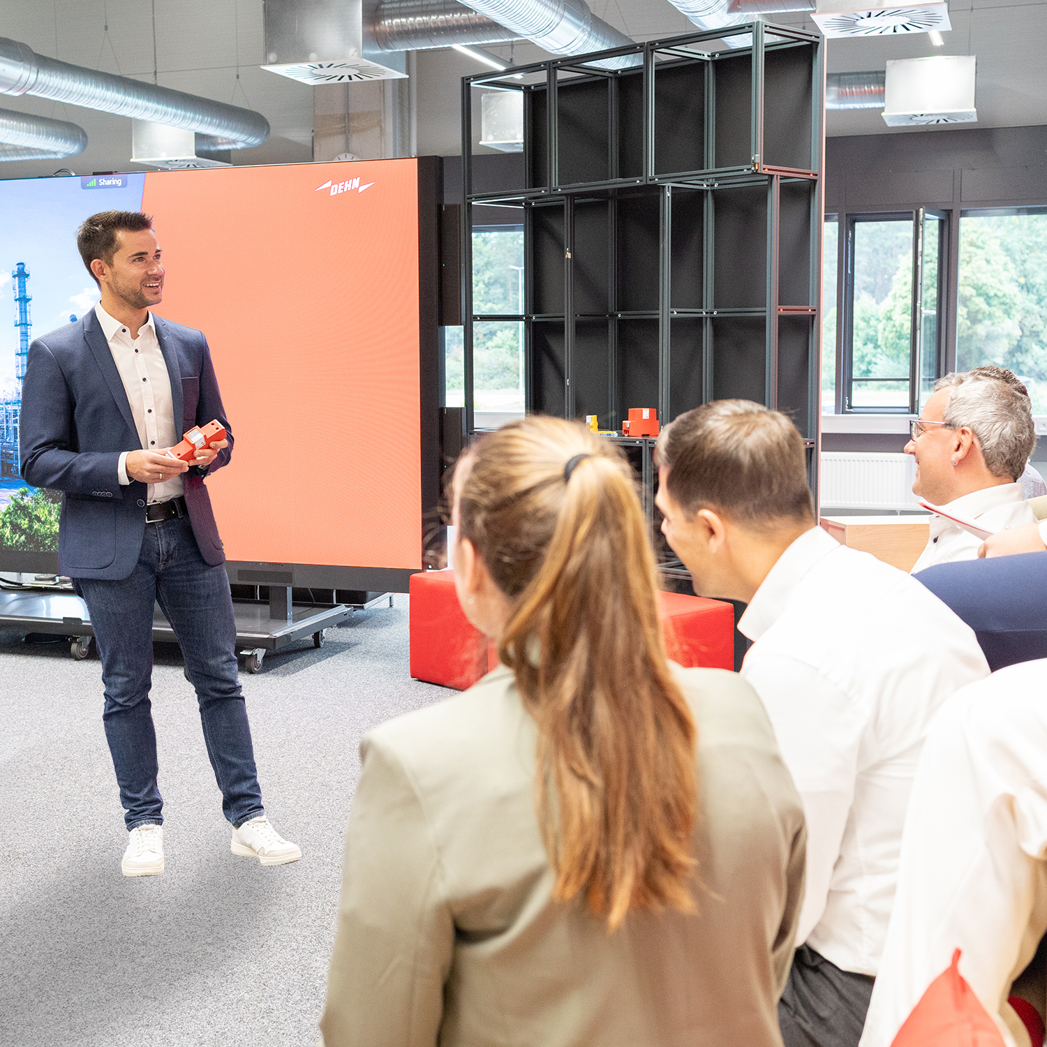 Man presenting a device to seated group in a room with large screen and industrial shelving.