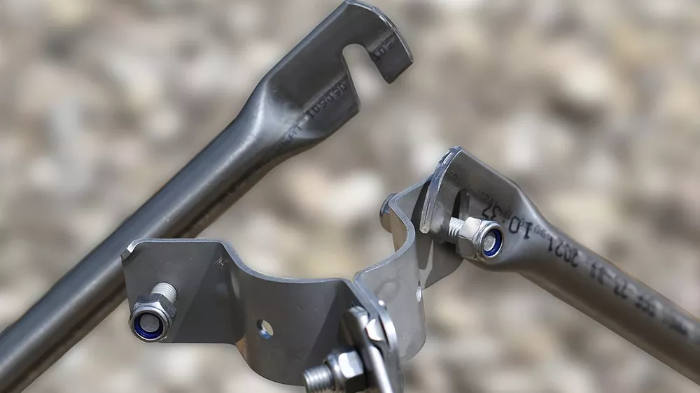 Close-up of metal rods connected by a clamp with bolts and nuts, showing open-ended joints on a blurred gravel background.