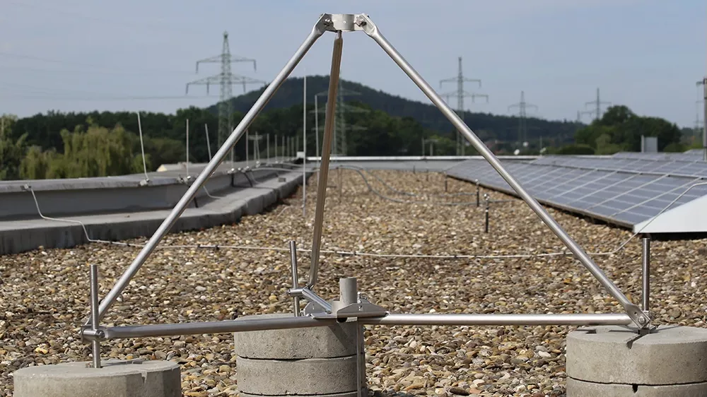Metal tripod structure mounted on concrete blocks on a gravel rooftop, with solar panels and power lines visible in the background.