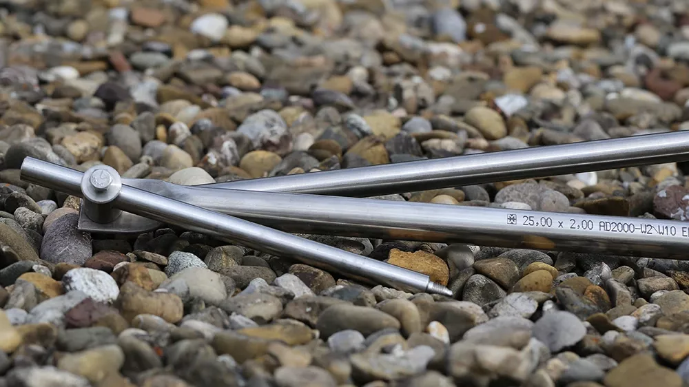 Metal rods with pointed ends lying on gravel, hinge joint visible, printed text “25,00 x 2,00 AD2000-UZ V10 E” on one rod.