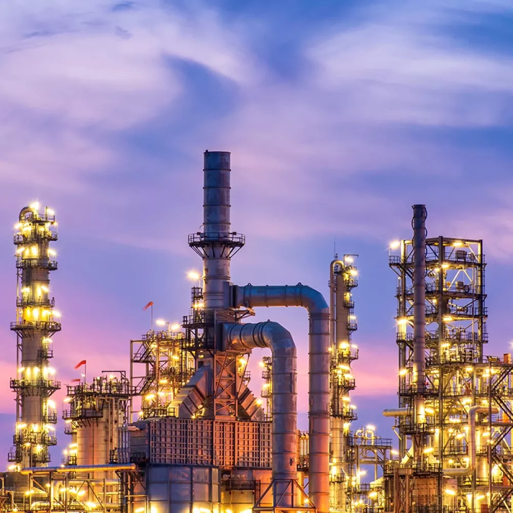 Beleuchtete Chemieanlage im Abendlicht mit zahlreichen Türmen, Pipelines und Lichtern vor einem lila gefärbten Himmel. 