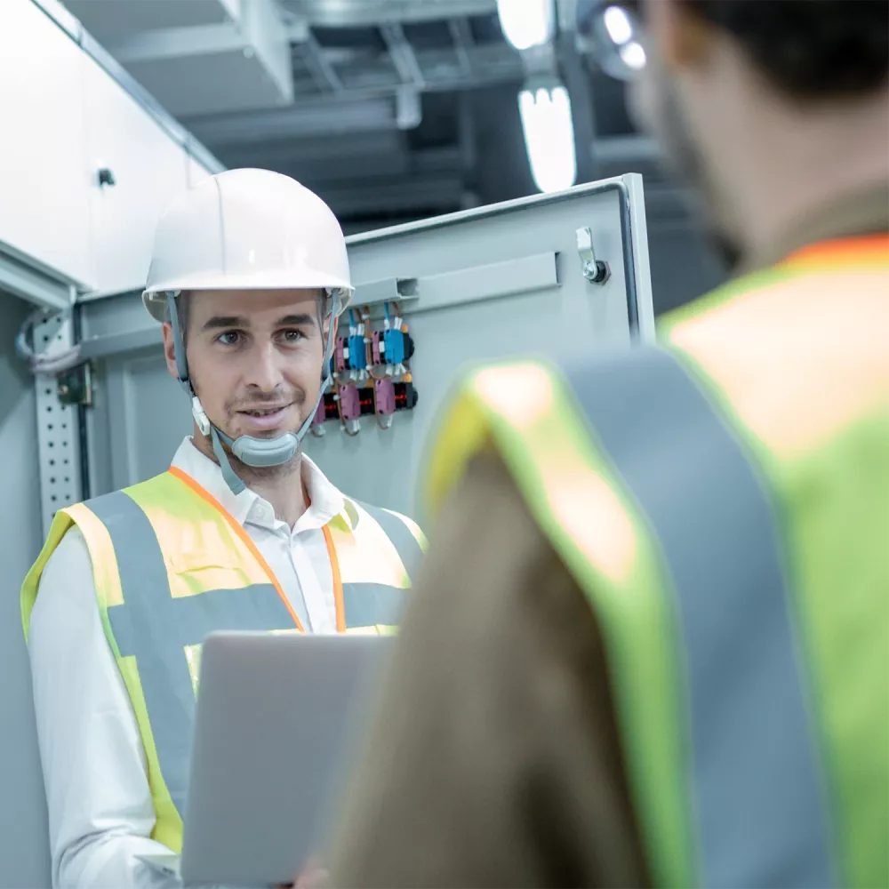 Engineer wearing a hard hat and high-visibility vest discusses an open electrical control cabinet with a colleague while holding a tablet in an industrial facility.