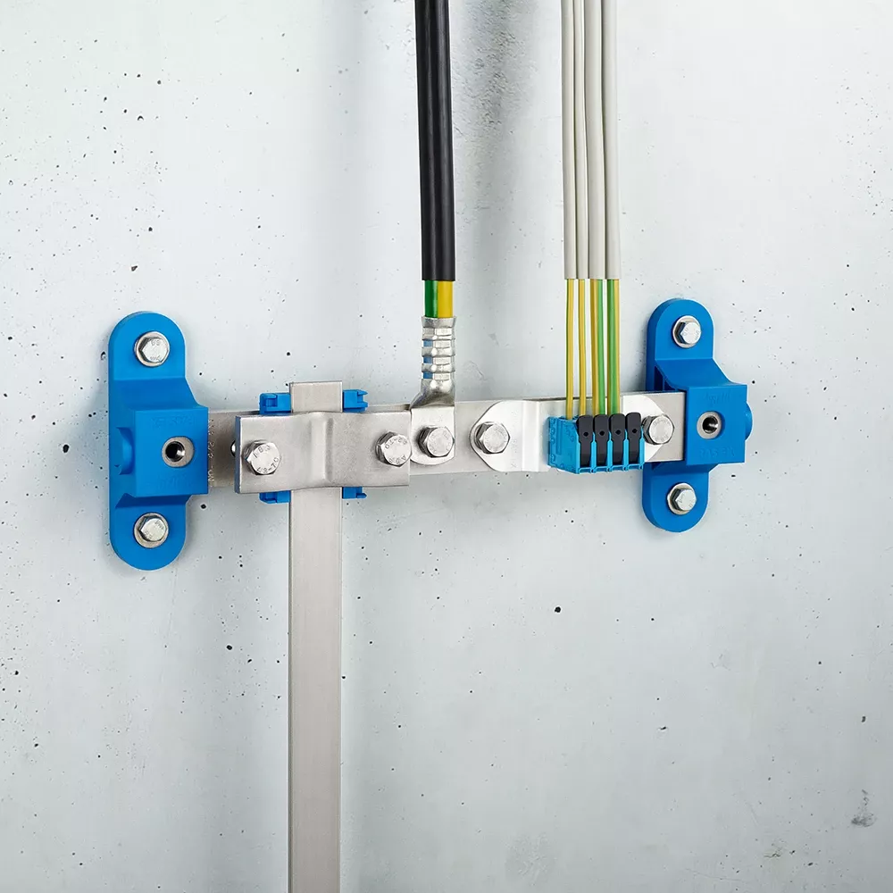 Wall-mounted grounding bar with metal clamps, connected cables, and blue mounting brackets on a light gray wall.