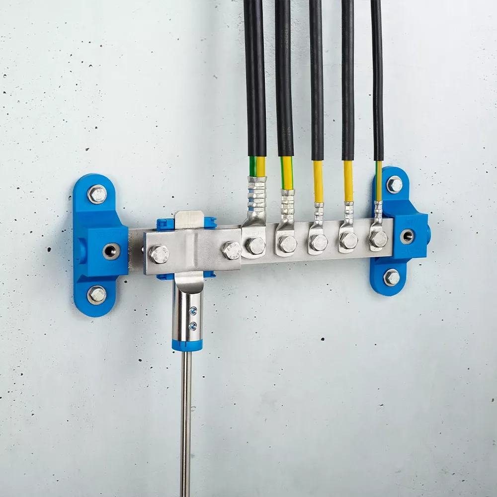 Wall-mounted grounding bar with metal clamps, connected black and yellow-green cables, and blue mounting brackets on a light gray wall.
