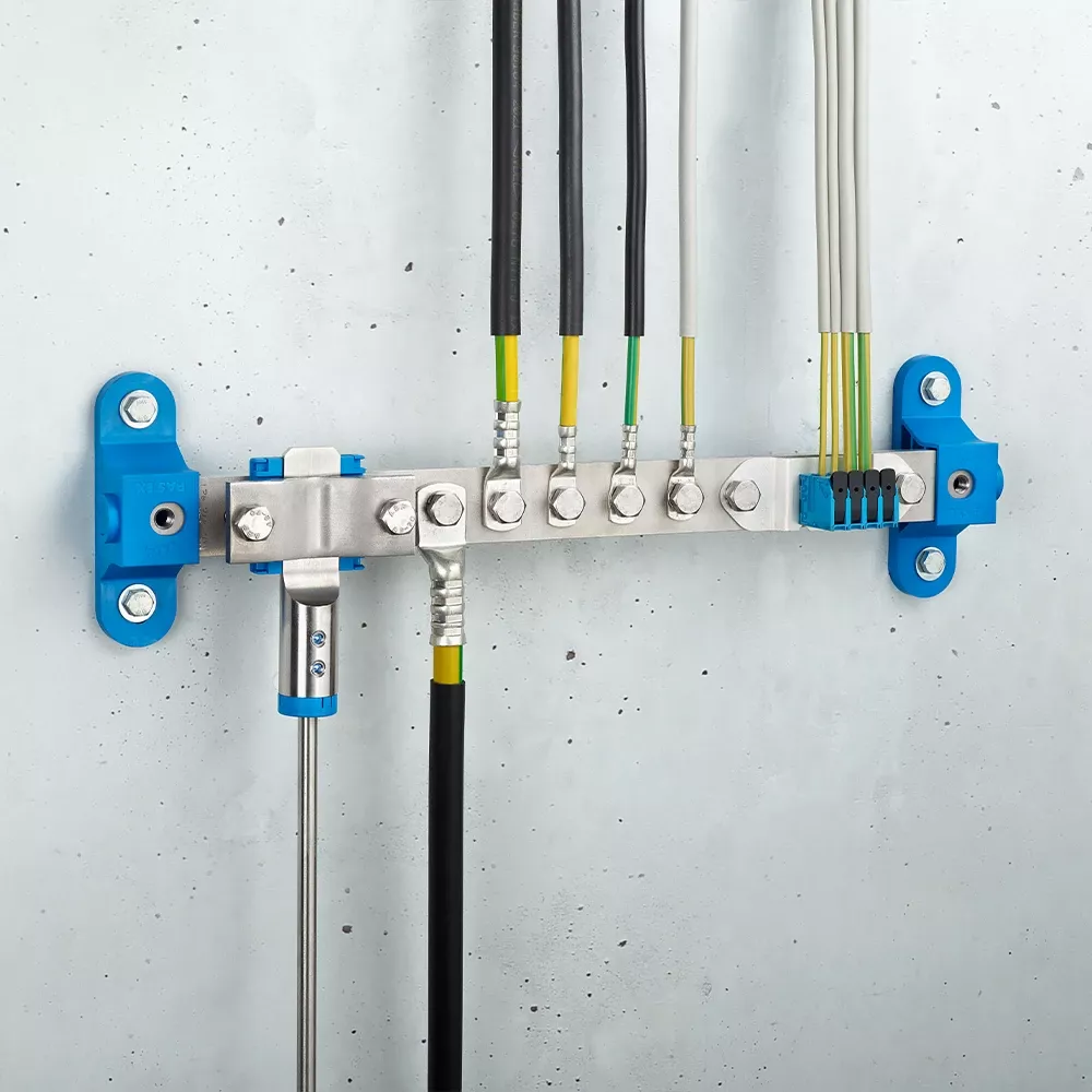 Wall-mounted grounding bar with metal terminals, connected black and yellow-green cables, and blue mounting brackets on a light gray wall.