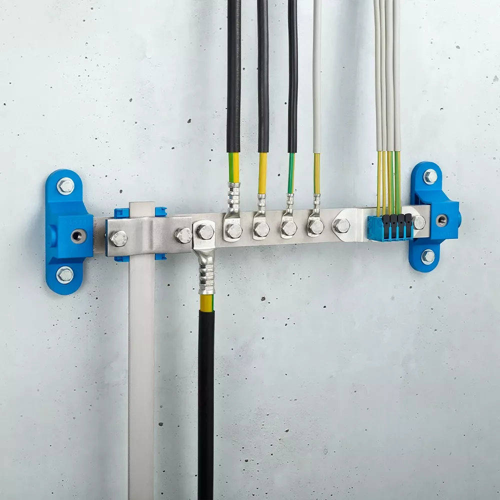 Wall-mounted grounding bar with metal terminals, connected black and yellow-green cables, and blue mounting brackets on a light gray wall.