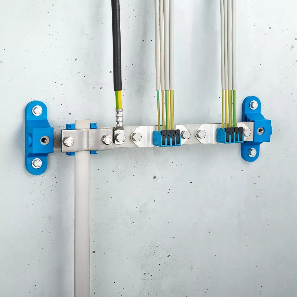 Wall-mounted grounding bar with metal terminals, connected black and yellow-green cables, and blue mounting brackets on a light gray wall.