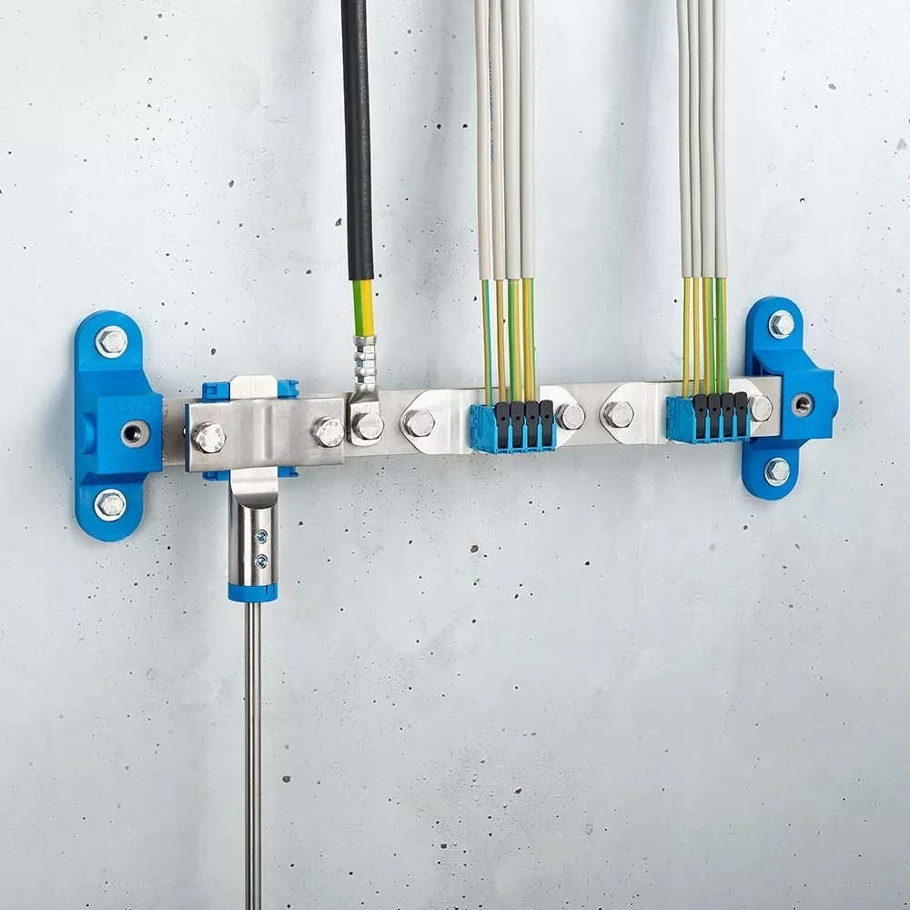 Wall-mounted grounding bar with metal clamps, connected black and yellow-green cables, and blue mounting brackets on a light gray wall.