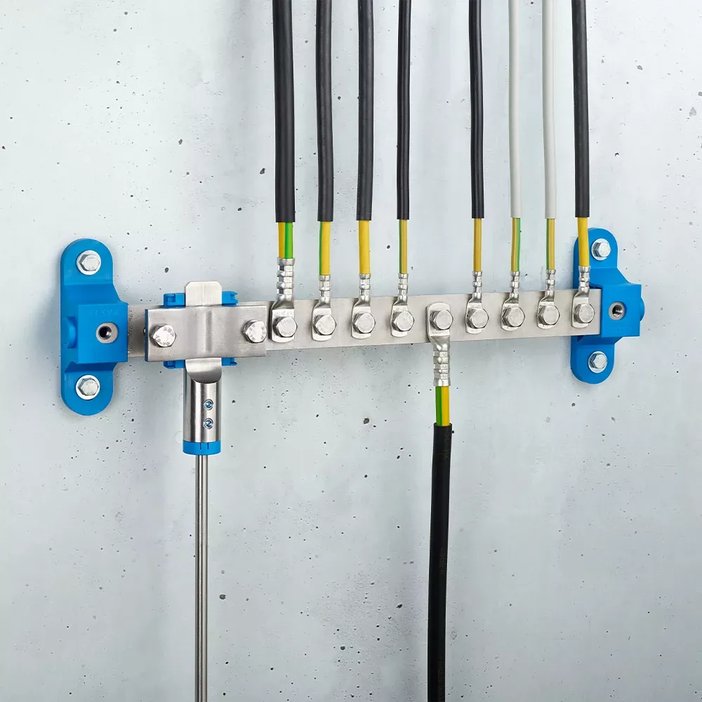 Wall-mounted grounding bar with metal terminals, connected black and yellow-green cables, and blue mounting brackets on a light gray wall.