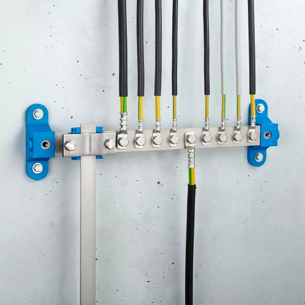 Wall-mounted grounding bar with metal terminals, connected black and yellow-green cables, and blue mounting brackets on a light gray wall.