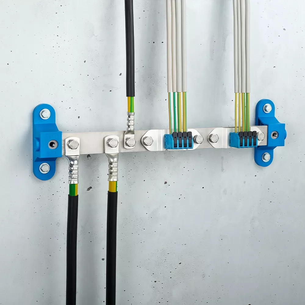 Wall-mounted grounding bar with metal terminals, connected black and yellow-green cables, and blue mounting brackets on a light gray wall.