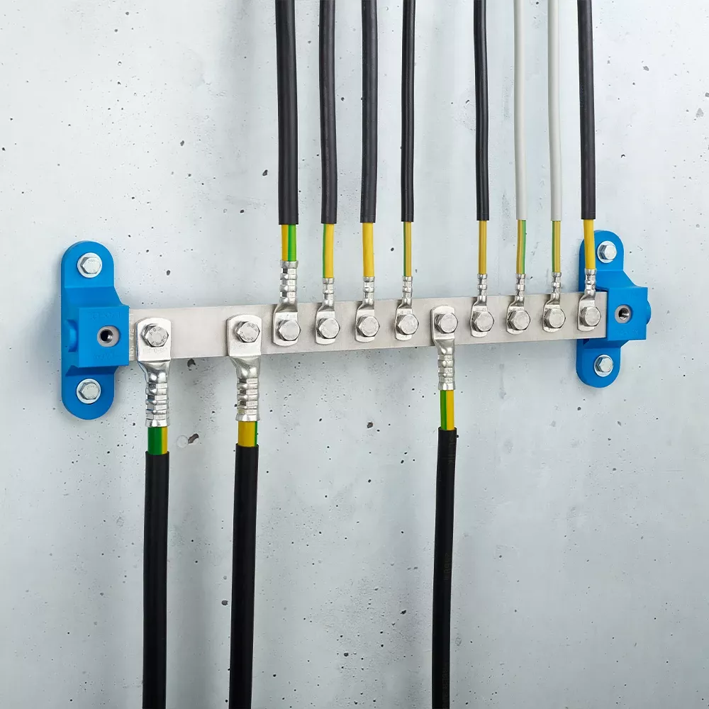 Wall-mounted grounding bar with metal terminals, connected black and yellow-green cables, and blue mounting brackets on a light gray wall.