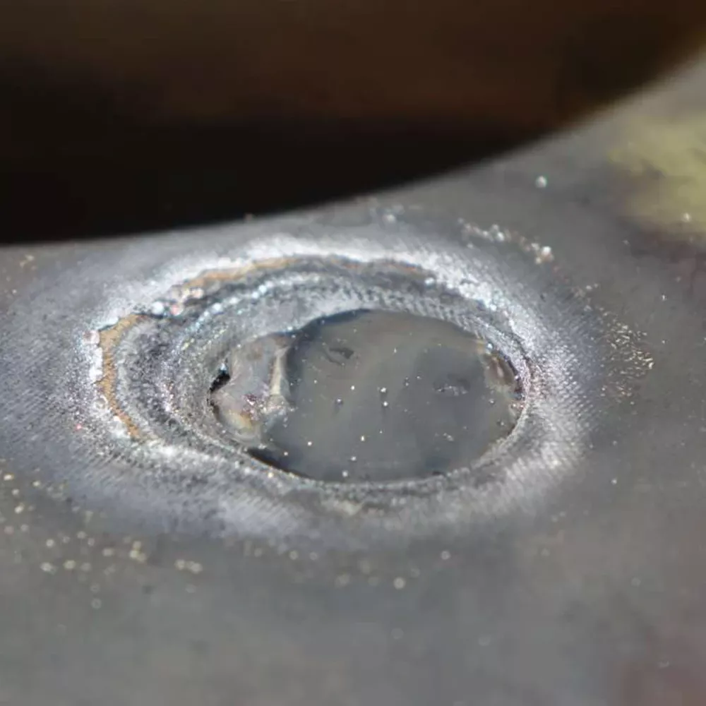Close-up of a circular weld spot on a metal surface with heat marks, rough edges, and small metal particles.