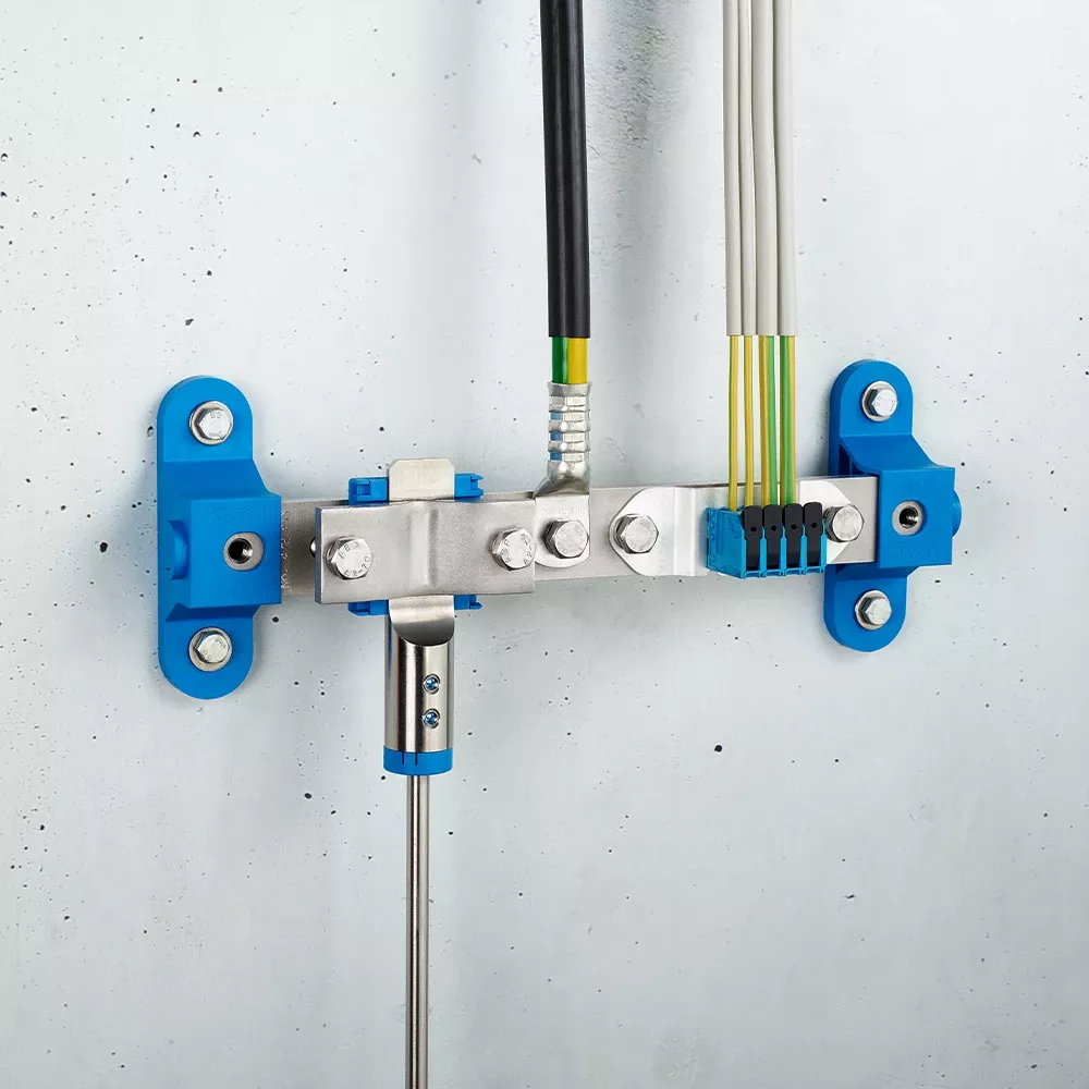 Wall-mounted grounding bar with metal clamps, connected black and yellow-green cables, and blue mounting brackets on a light gray wall.