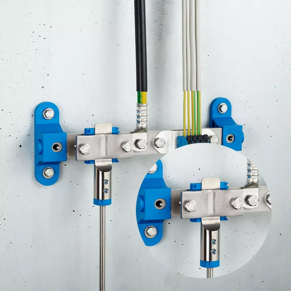 Metal grounding bar on a wall with connected black and white cables, green-yellow conductors, blue mounting brackets, and a metal pipe clamp.