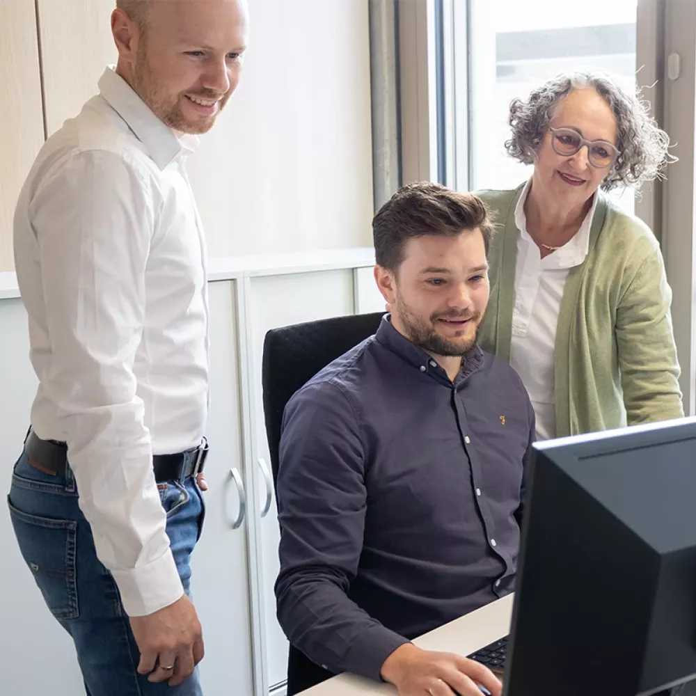 Three people at a desk in an office, two standing and one seated, looking at a computer monitor together near a window.