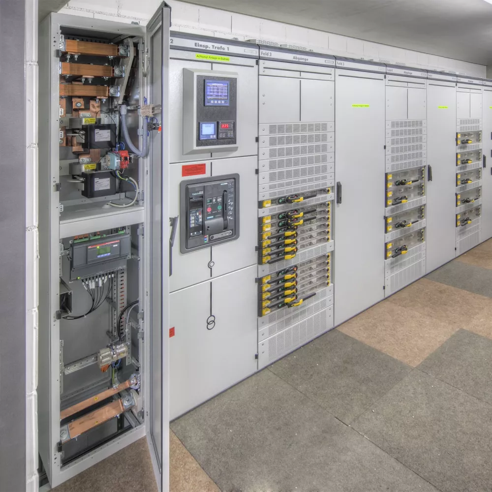 Large electrical switchgear cabinet room with open control panel showing busbars, circuit breakers, and monitoring displays.