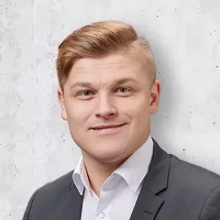 Portrait of a young man in a dark suit jacket and white shirt, smiling slightly against a light gray background.