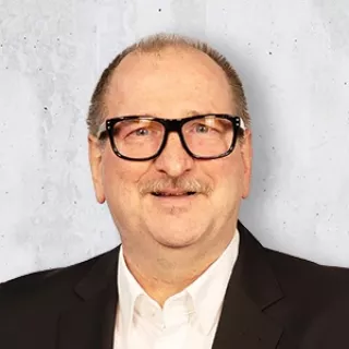 Portrait of a man wearing black-framed glasses, a dark suit jacket and white shirt, smiling against a light gray background.